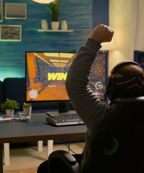 Videogamer player raising hands after winning first person shooter competition wearing hradphones. Professional pro gamer playing online video games with new graphics on powerful computer